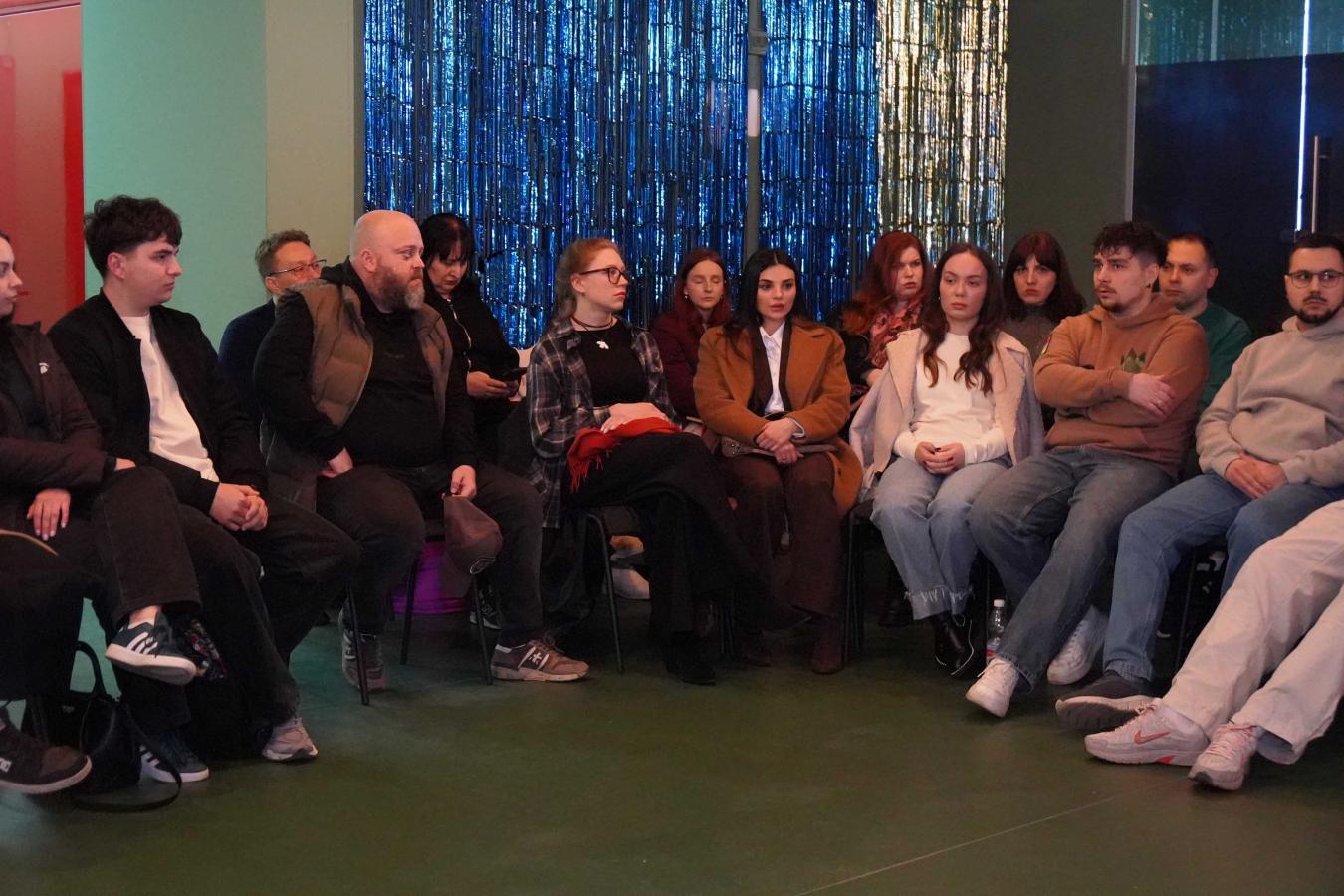  Близько п'ятнадцяти осіб різного віку (переважно молодь) зосереджено слухають або спостерігають за чимось поза кадром. Вони одягнені в повсякденний одяг: худі, джинси, куртки та светри.