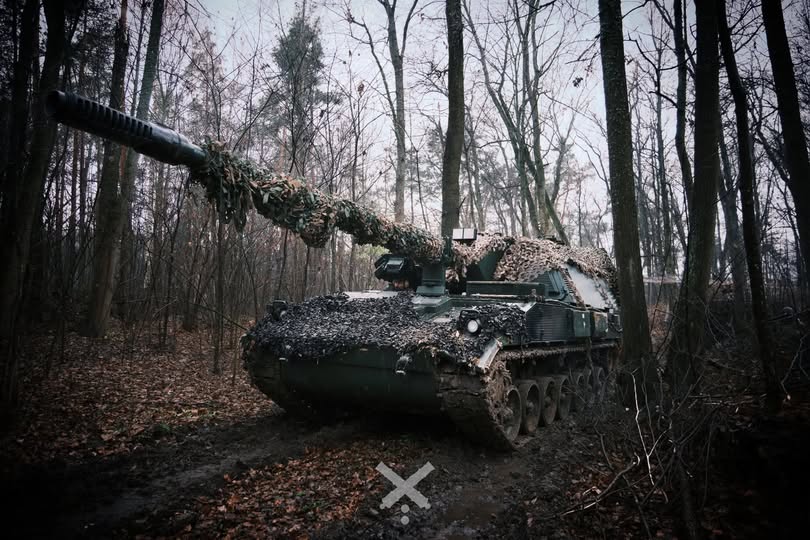 На фото серед зимового лісу стоїть гусенична самохідна артилерійська установка темного кольору, замаскована сітками й гіллям по всьому корпусу та вздовж довгого ствола гармати. Машина розташована на вкритій листям ґрунтовій дорозі між деревами, навколо — похмуре небо та голі стовбури, що підкреслюють приховану бойову позицію техніки