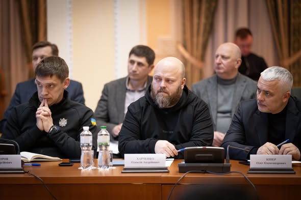 На фотографії зображено групу чоловіків, які сидять за столом під час офіційної наради, при цьому на ідентифікаційних картках видно імена «Микола Якович Нарижний» (центральний чоловік з бородою) та «Василь Іванович Книга» (чоловік праворуч у чорному піджаку)