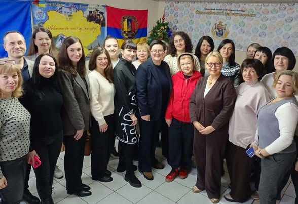 Групове фото учасників заходу соціальної сфери Луганщини в укритті: декілька десятків людей стоять щільним колом у приміщенні, на задньому плані – стінди з логотипами, прапорами та мапою України, поруч новорічна ялинка, усі одягнені в повсякденний одяг і дивляться в бік камери