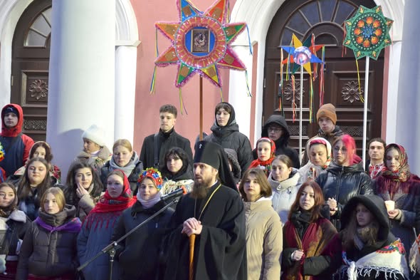 Група людей різного віку, серед яких багато молоді в національному українському одязі та зимових куртках, зібралася на вулиці перед будівлею з білими колонами та арковими дверима. У центрі композиції — чоловік у чорному церковному вбранні та головному уборі, схожому на священника. Деякі учасники тримають великі, багато прикрашені різдвяні зірки-вертепи, що є традиційним атрибутом українського різдвяного дійства — вертепу