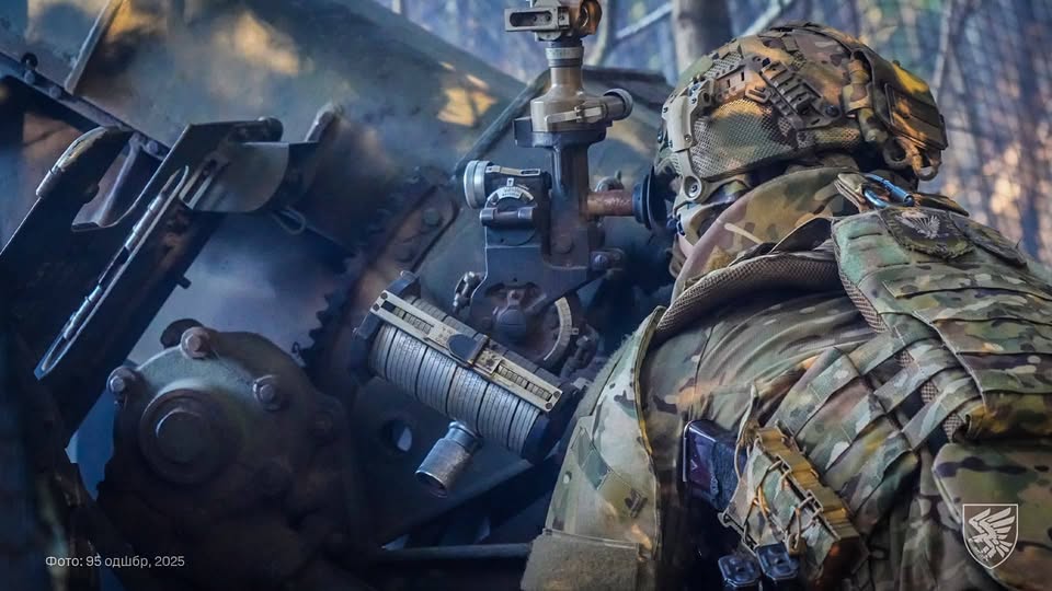 Український артилерист готується до чергового пострілу, уважно звіряючи приціл на позиції ворога в лісосмузі. Серед ретельно замаскованих гармат і техніки він зосереджено працює з оптичним приладом, перетворюючи координати розвідки на точний вогонь. Захисник у сучасному камуфляжі й шоломі символізує витривалість і професіоналізм української армії, що тримає оборону на передовій