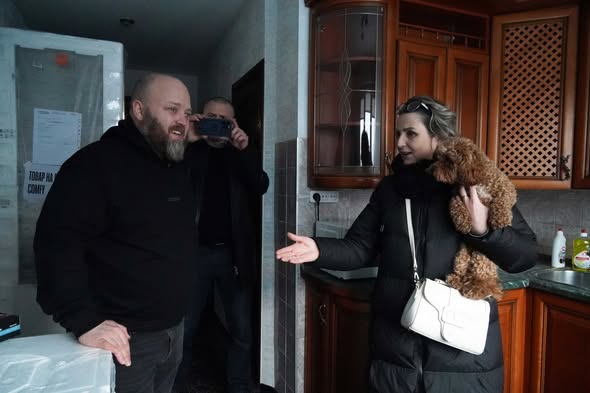 На фотографії зображено людей у приміщенні, схожому на кухню, де жінка тримає на руках коричневого пуделя, а поруч стоїть чоловік у чорній кофті, тоді як інший чоловік на задньому плані знімає сцену на камеру
