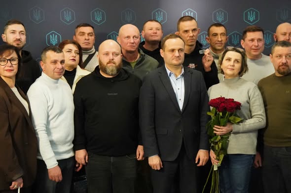 Групове фото колективу робочої команди, ймовірно, з Координаційного центру допомоги ВПО у Кропивницькому. На темному фоні з повторюваними логотипами тризуба та написом "НА" (можливо, "НАШ" чи "НАЦІЯ") позують понад 20 осіб різного віку, переважно чоловіків середнього віку з короткими зачісками та бородами, а також кілька жінок. Центральна постать — чоловік у темному костюмі з синьою сорочкою тримає букет червоних троянд, інші одягнені в повсякденний одяг: светри, куртки, худі