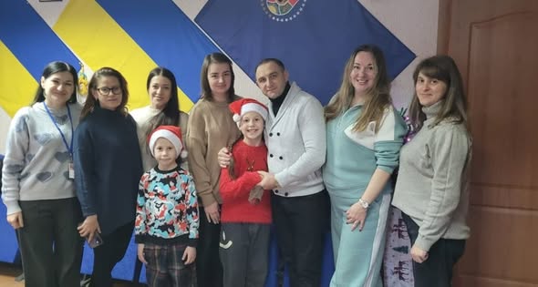 У приміщенні на фоні великого синьо-жовтого прапора та ще одного синього прапора з емблемою стоїть група дорослих і двоє дітей у новорічних шапках. Усі позують щільним колом, деякі тримають дітей за плечі чи за руки, створюючи атмосферу дружньої святкової зустрічі.
