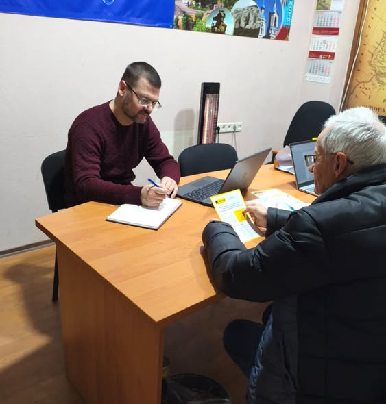 Наданий альтернативний опис до фото: два чоловіки сидять за офісним столом, один з яких пише в блокноті, а інший розглядає документи
