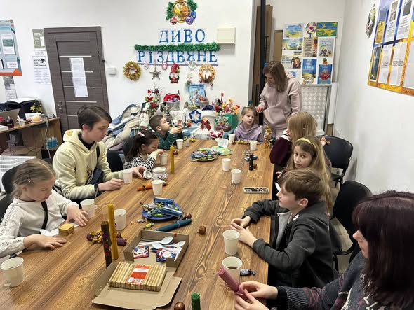 кімната з двова великим вікнами, посред кімнати стоїть великий дерев'яний стіл, за яким зібралося багато дітвори. На столі стоять смаколики, різдвяні свічки.
