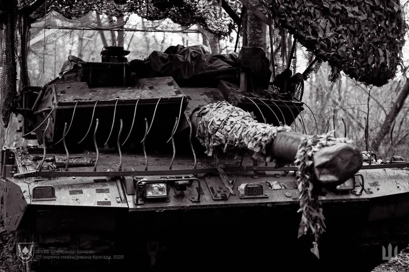На чорно-білій фотографії зображено передню частину танка, ймовірно Stridsvagn 122 або Leopard 2, який належить 121-й окремій механізованій бригаді Збройних Сил України, оснащений додатковим динамічним захистом та камуфляжем, і розташований у лісовій місцевості