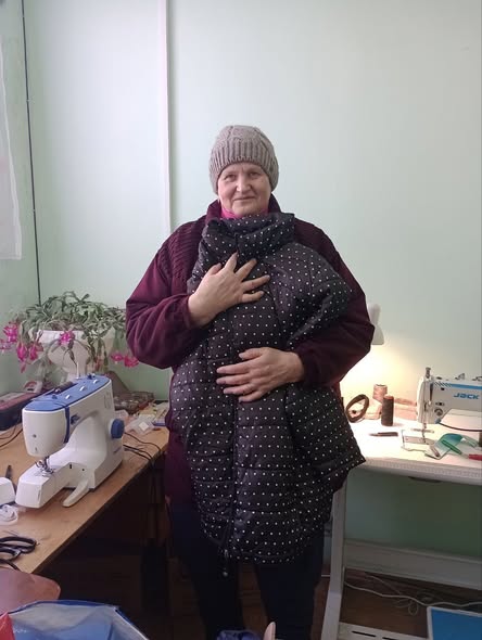 На фотографії зображено літню жінку в'язаній шапці та бордовому светрі, яка стоїть у приміщенні з двома швейними машинками на столах і з посмішкою обіймає пухку дитячу або домашню тварину куртку чи конверт-спальник темно-сірого кольору в білий горошок, що свідчить про діяльність, пов'язану з шиттям або благодійністю.