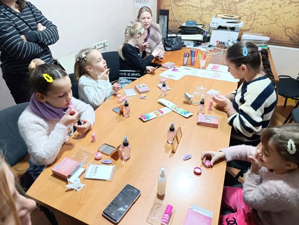 Група дітей та дорослих бере участь у майстер-класі з косметики та догляду за собою, сидячи за офісним столом, заповненим різноманітними продуктами та інструкціями