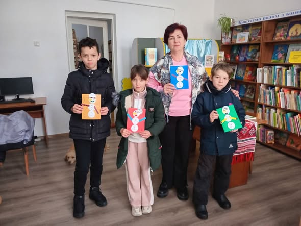 Чотири дитини та доросла жінка, ймовірно, вчителька чи бібліотекарка, стоять разом у бібліотеці та гордо демонструють свої зимові вироби-аплікації із зображеннями сніговиків. Цей альтернативний текст описує основну сцену, включаючи кількість людей, місце дії та предмети, які вони тримають.