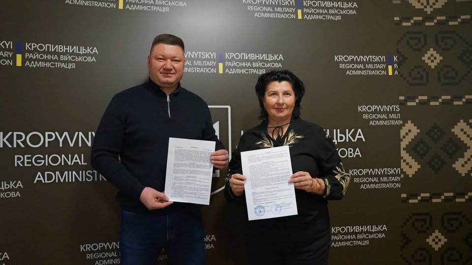 Двоє людей, чоловік та жінка, позують для офіційного фото в приміщенні з брендуванням Кропивницької районної військової адміністрації, тримаючи в руках підписані документи