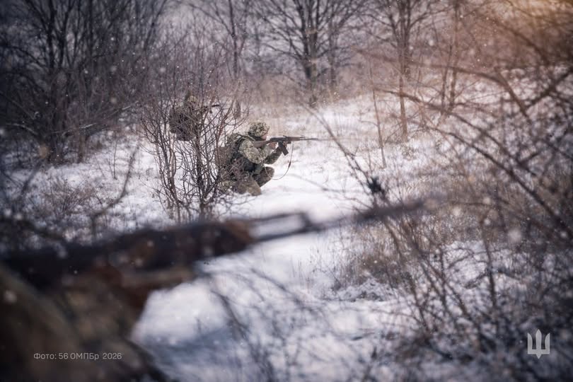 На фотографії зображено українського військовослужбовця, який перебуває на бойовій позиції в сніговому лісі або полі, цілиться з автомата та ховається серед гілок, що свідчить про виконання бойового завдання в зимових умовах.
