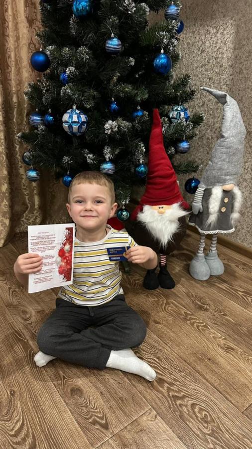 Щасливий маленький хлопчик сидить на дерев'яній підлозі перед різдвяною ялинкою, прикрашеною синіми кульками, та двома декоративними гномами, гордо демонструючи білу листівку та синю картку, які він тримає в руках