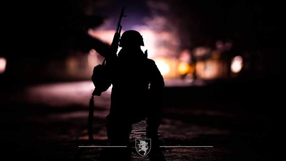 на темному тлі, освітленому лише розмитими вогнями міста або військової бази, зображено силует озброєного солдата, який стоїть на коліні з автоматом, а внизу розташований герб або емблема військової частини, що підкреслює урочистість моменту