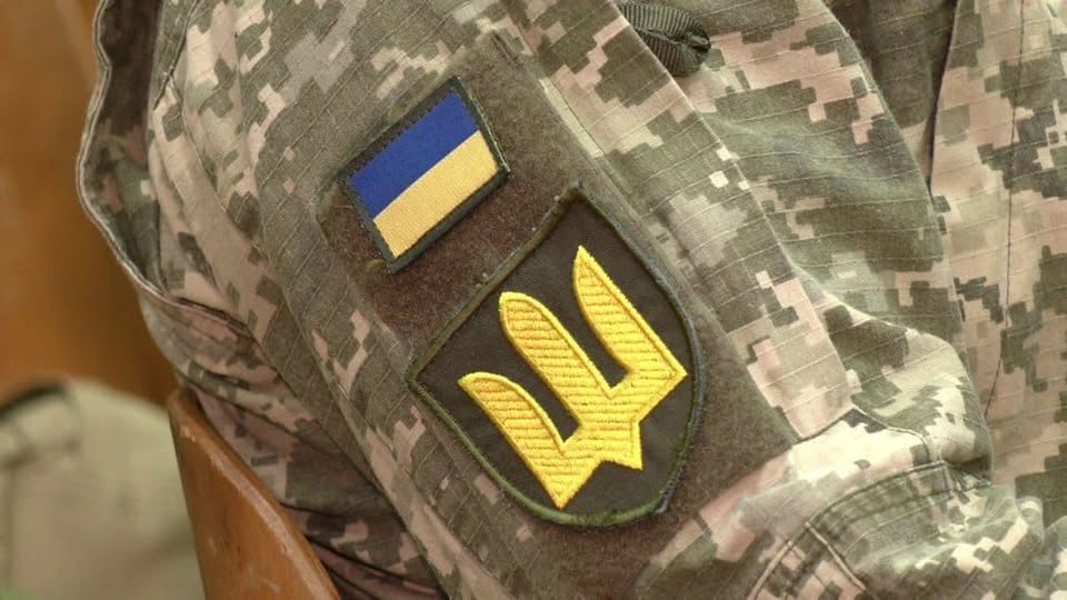 Крупним планом зображено рукав військової форми зі сучасним піксельним камуфляжем, на якому розташовані дві нашивки: менша прямокутна із синьо-жовтим прапором України та більша емблема тризуба золотистого кольору на темному щиті