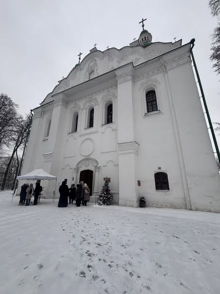 Біла церква з зеленим куполом стоїть на засніженій землі, де група людей зібралася біля входу поруч із маленькою прикрашеною ялинкою та білим наметом.