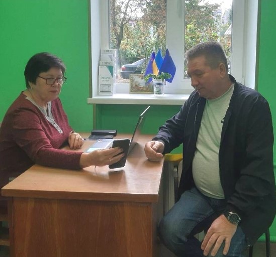 жінка в окулярах та чоловік у куртці, сидять за дерев'яним столом у приміщенні з яскраво-зеленими стінами та обговорюють щось, дивлячись на смартфон жінки, тоді як на підвіконні видніються маленькі українські прапори, що свідчить про офіційну або соціальну зустріч в Україні.