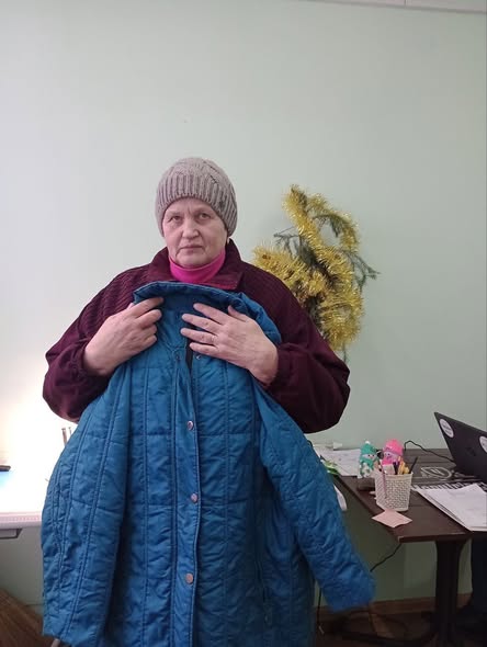 жінка середнього віку в бордовій куртці та сірій в'язаній шапці стоїть у приміщенні зі світло-зеленими стінами, тримаючи перед собою синій стьобаний плащ, а на задньому плані видніється декоративна ялинка та робочий стіл
