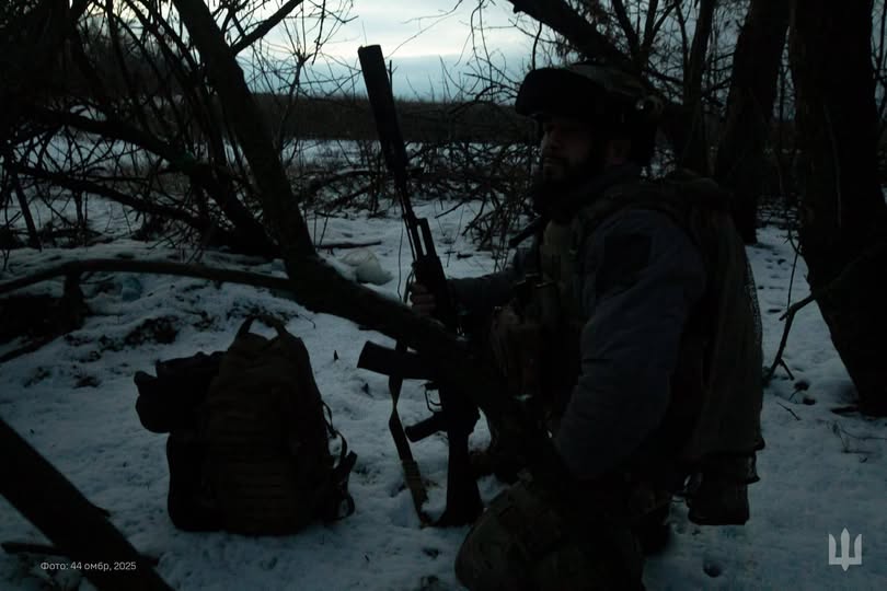 Український військовослужбовець у повному бойовому спорядженні, включаючи шолом, тактичний жилет і рюкзак, сидить на колінах на засніженій землі в густих заростях, тримаючи в руках автоматичну зброю з прикріпленим великим глушником, на тлі темного зимового пейзажу