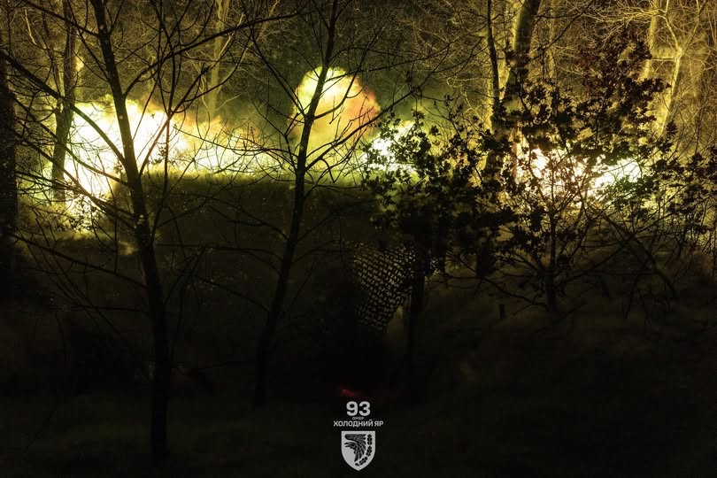 Темна нічна сцена в лісі із силуетами голих дерев і вічнозелених кущів, освітлена яскравим жовтим світлом на задньому плані, із нашивкою 93-ї бригади «Холодний Яр» унизу по центру