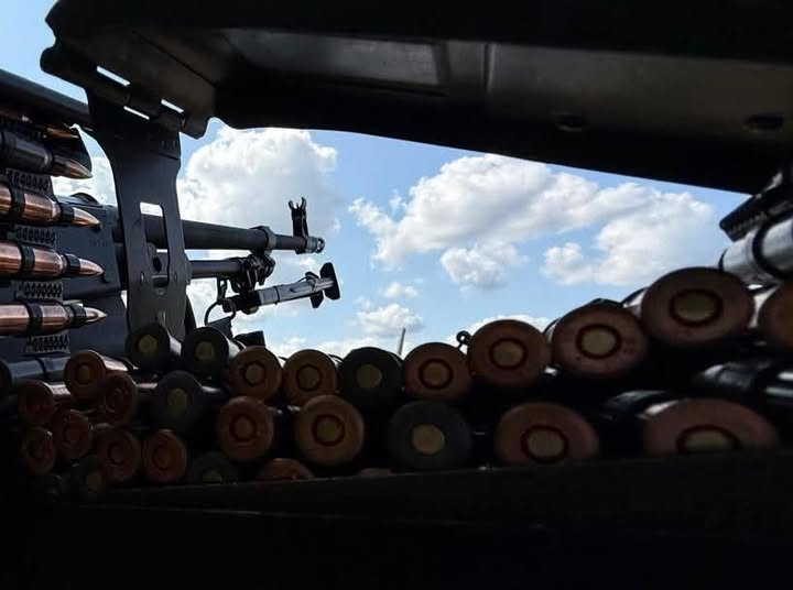 Вид ізсередини бойової позиції або військової техніки, що демонструє великокаліберний кулемет та численні боєприпаси, спрямовані на тло блакитного неба з хмарами