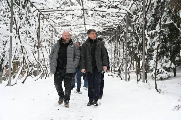 Два чоловіки в зимовому одязі йдуть засніженим проходом, утвореним аркою з покритих снігом гілок, із додатковими людьми та деревами на задньому плані.