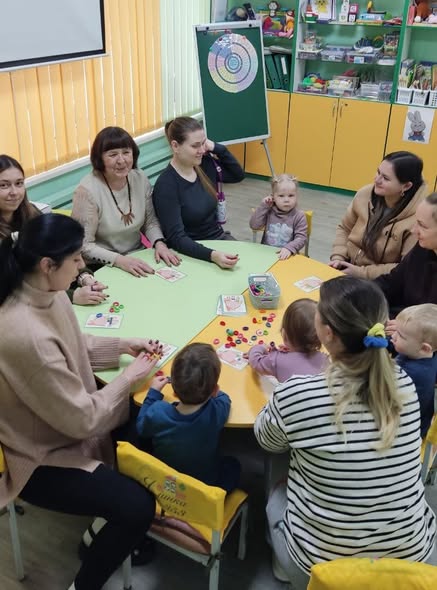 Група дорослих і дітей сидить навколо зеленого столу в класі або дитячому закладі, де розкладені іграшки або навчальні матеріали для спільної гри