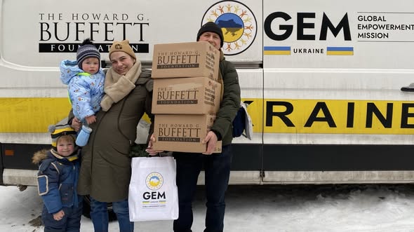 українська сім'я з двома дітьми стоїть на засніженій землі біля вантажівки з логотипами Howard G. Buffett Foundation та GEM Ukraine, отримуючи пакунки гуманітарної допомоги від цих благодійних організацій