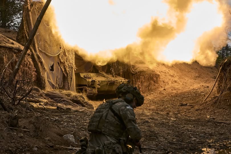 український солдат на передньому плані, який спостерігає, як бронетранспортер стріляє, створюючи яскравий вибух і клуби диму з вогнем у прихованій вогневій позиції, ймовірно, під час російсько-української війни.