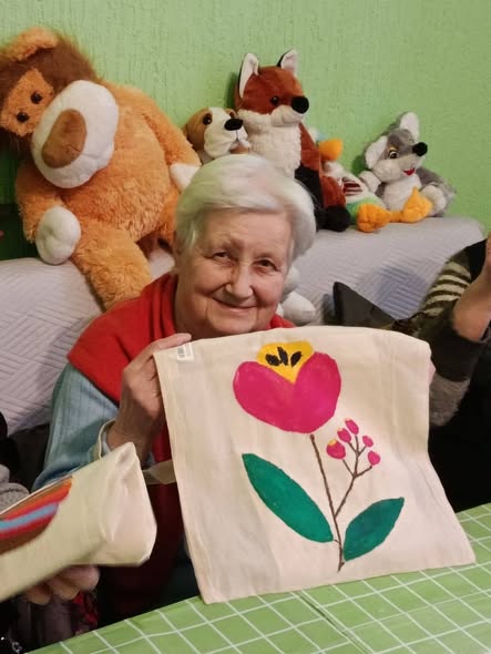 На зображенні літня жінка з сивим волоссям посміхається, сидячи серед м'яких іграшок, і тримає світлу тканину з малюнком червоного тюльпана та зелених гілочок