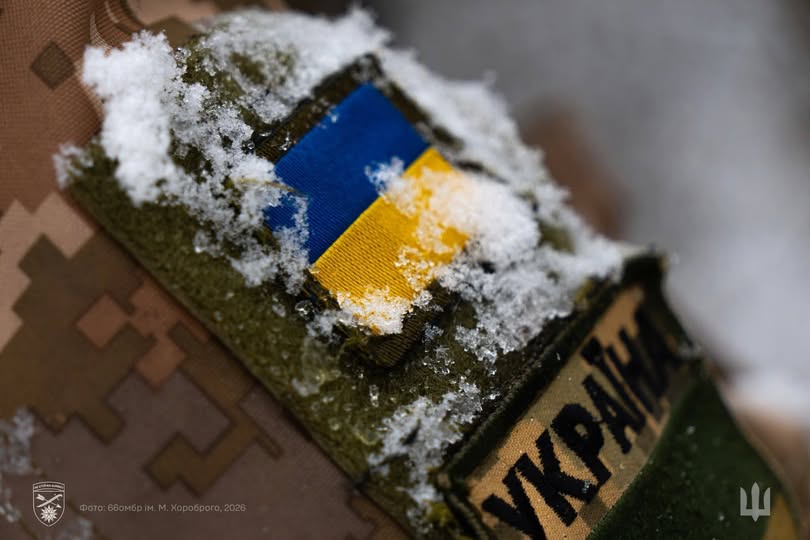 Крупний план військової форми з нашивкою українського прапора та написом «УКРАЇНА», присипаних снігом, з додатковим логотипом та текстом дати в нижньому лівому куті.
