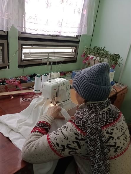Людина в синій в'язаній шапці та светрі з норвезьким візерунком сидить за дерев'яним столом і шиє на білій швейній машині білу тканину, з додатковими нитками та рослинами на фоні