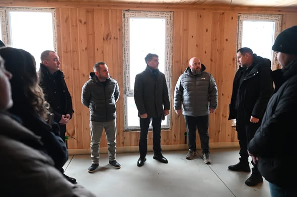 Кілька чоловіків у зимовому одязі стоять у кімнаті з дерев’яними стінами, обговорюючи будівництво житлового комплексу для внутрішньо переміщених осіб на Закарпатті. Вони, зокрема, включають голову Луганської обласної військової адміністрації та голову Закарпатської обласної державної адміністрації Мирослава Білецького.