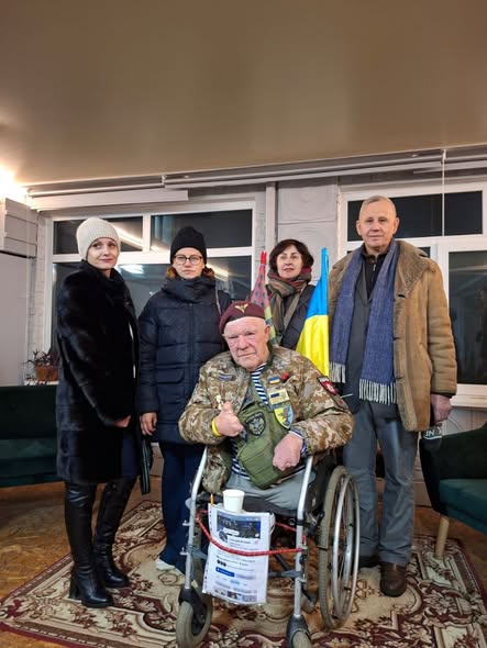 зображено українського волонтера дядька Гришу (Григорія Янченка) в інвалідному візку, який сидить у приміщенні з групою інших людей, зокрема жінками у зимовому одязі та чоловіком у пальті, тримаючий невеликий синьо-жовтий прапор