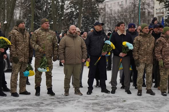 Група чоловіків у військовій формі стоять на вулиці взимку, деякі тримають букети синьо-жовтих квітів.