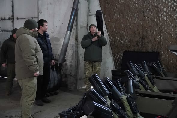 На зображенні група з трьох чоловіків у зимовому та військовому одязі перебуває в приміщенні, схожому на склад або майстерню. На передньому плані видно кілька рядів чорних ракетних або протитанкових комплексів, акуратно складених у стелажах.