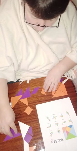 Зображення показує людину в окулярах, яка сидить за дерев'яним столом і складає фігури з оранжевих та фіолетових частин головоломки танграм, поруч лежить аркуш із прикладами та написом TANGRAM