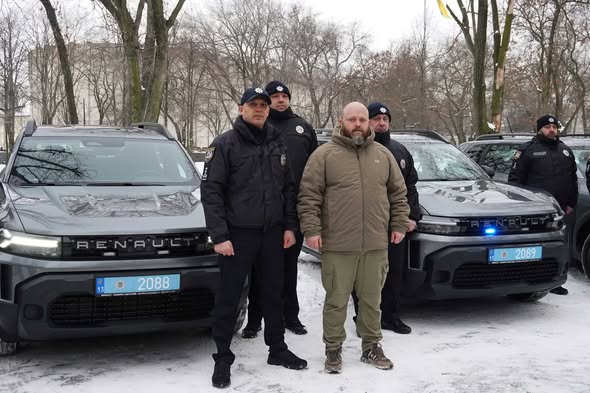 Група чоловіків у поліцейській формі та цивільному одязі, включно з Оксеном Лісовим (другий праворуч), стоять біля нових службових автомобілів Renault Duster. Ці автомобілі (з номерними знаками, що містять цифри 2088 та 2089) були передані підрозділам Національної поліції України, зокрема для поліцейських офіцерів громад. Подібні передачі автомобілів регулярно відбуваються в різних регіонах України, включаючи прифронтові області, для покращення матеріально-технічного забезпечення правоохоронців.