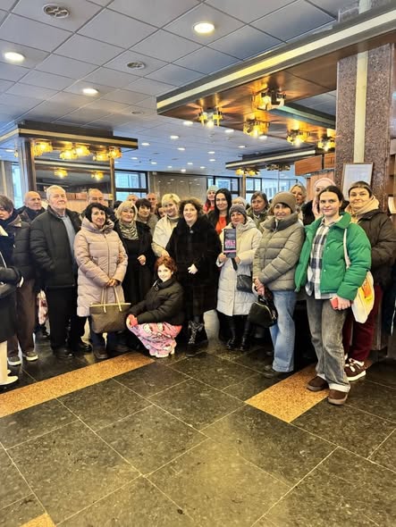 Група з близько двох десятків людей різного віку в зимовому одязі зібралася в приміщенні з темною плиткою на підлозі та декоративними світильниками на стелі