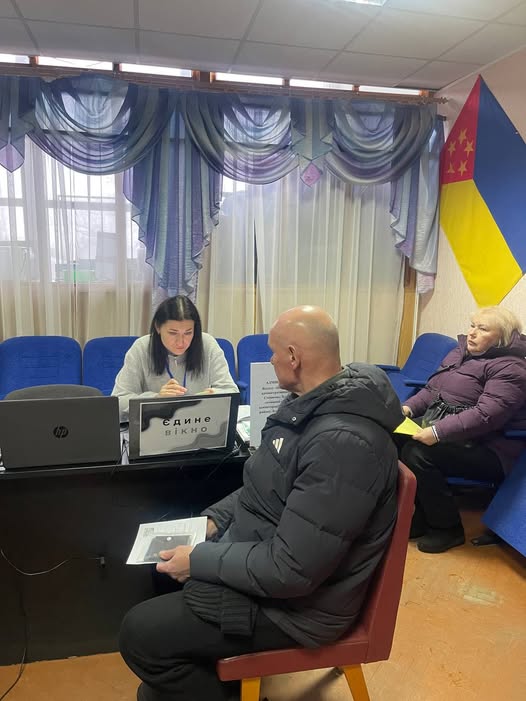 На передньому плані сидить лисий чоловік у темній куртці, який тримає документ і чекає. За столом праворуч від нього сидить жінка за ноутбуком, перед якою стоїть табличка з написом «ЄДИНЕ ВІКНО». Праворуч на синьому стільці сидить літня жінка у фіолетовій куртці. На задньому плані видно вікно із синіми шторами та на стіні — частину українського прапора та прапора іншого регіону