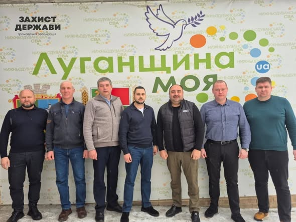 На зображенні група з шести чоловіків стоїть у приміщенні перед великим рекламним банером. На банері написано «Луганщина МОЯ» та «ЗАХИСТ ДЕРЖАВИ», а також зображено голуба миру та різнокольорові кола. Чоловіки одягнені у повсякденний одяг, включаючи светри, джинси та жилети