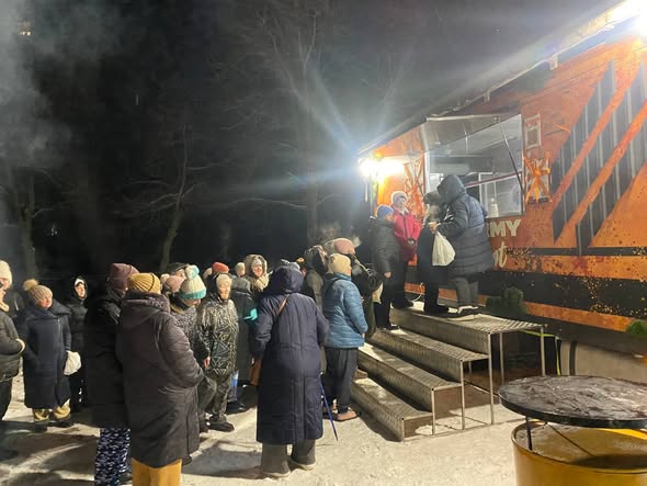 На зображенні показана група людей на вулиці вночі, які стоять біля великого оранжевого автомобіля, можливо, фудтрака, на снігу. Люди одягнені в зимовий одяг.