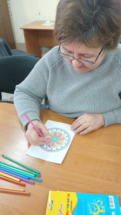 Людина в окулярах і сірому светрі сидить за дерев'яним столом і розфарбовує малюнок мандали рожевим олівцем. На столі видно розсипані кольорові олівці та відкриту упаковку олівців українського бренду «Барвінок» на 12 кольорів
