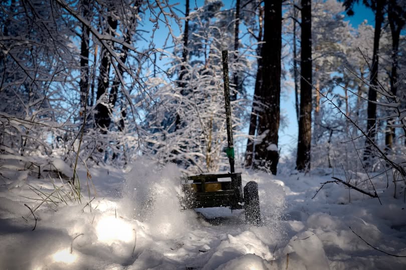На зображенні показано невеликий військовий наземний роботизований комплекс (НРК) на колесах зеленого кольору, який активно рухається глибоким снігом у засніженому лісі, створюючи бризки снігу. Такі роботи використовуються українськими військовими для різних завдань, включаючи розвідку та евакуацію