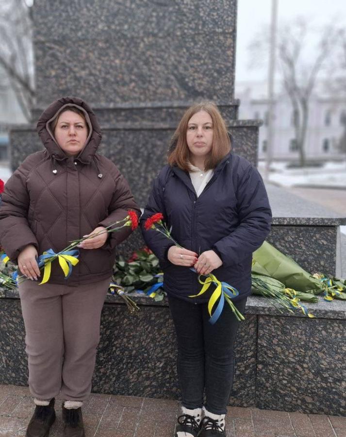 Дві жінки стоять поруч із гранітним пам'ятником або меморіалом у засніженому парку взимку. Вони тримають букети квітів (деякі червоні гвоздики, деякі жовто-сині квіти), перев'язані синьо-жовтими стрічками, що символізують український прапор. На землі біля основи пам'ятника також лежать інші букети квітів та стрічки.