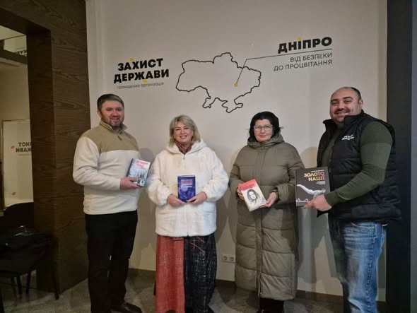 На зображенні четверо людей, троє з яких тримають книги, позують у приміщенні біля стіни з картою України та написами українською мовою: «ЗАХИСТ ДЕРЖАВИ», «ДНІПРО», «ВІД БЕЗПЕКИ ДО ПРОЦВІТАННЯ» та «ЗОЛОТО НАШ». Люди посміхаються і одягнені в повсякденний одяг, один із чоловіків носить жилет