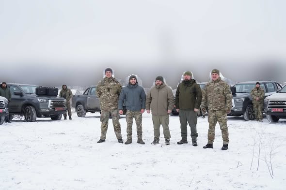На зображенні група з п'яти чоловіків у військовій формі та зимовому одязі стоїть на засніженому полі перед кількома пікапами сірого кольору. Також на задньому плані видно ще кількох людей і транспортні засоби