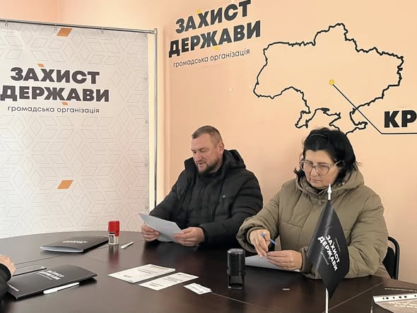 На зображенні двоє людей, чоловік і жінка в зимовому одязі, сидять за столом у кімнаті. Вони тримають папери, а на столі перед ними лежить інша документація, ручка, печатка та невеличкий чорний прапорець з написом «ЗАХИСТ ДЕРЖАВИ». На стіні позаду них розташовані банери з великими написами «ЗАХИСТ ДЕРЖАВИ» та контуром карти України