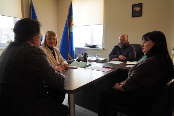 Зображення є фотографією, зробленою в офісному приміщенні, де за великим дерев'яним столом сидять чотири людини, що проводять зустріч або нараду